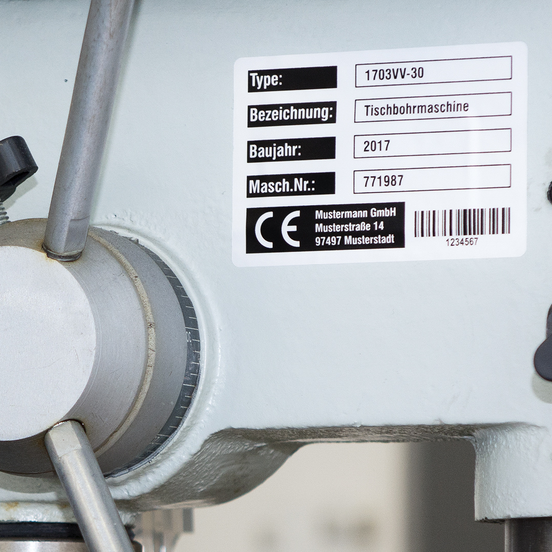 Polyester Typenschildetikett mit Maschinendaten, CE-Zeichen und Barcode auf einer grauen Tischbohrmaschine &ndash; langlebige Kennzeichnung f&uuml;r industrielle Anwendungen.