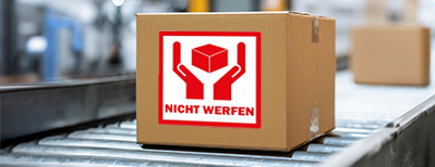 Karton mit rotem Warnaufkleber 'Nicht werfen' auf einem Förderband in einer Versandhalle.