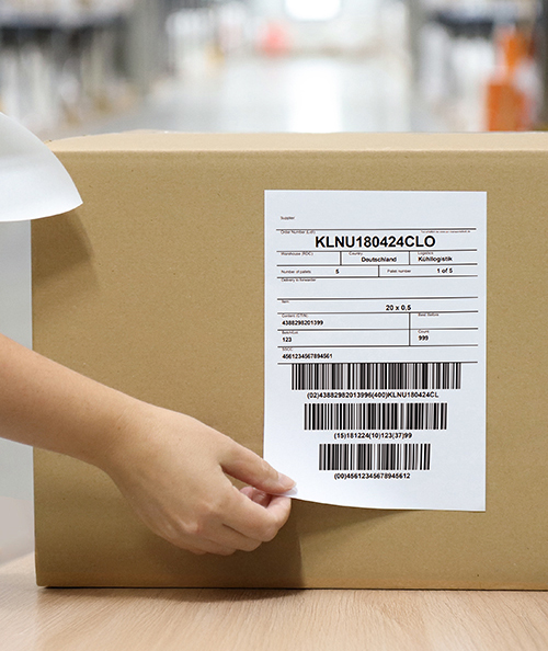 Versandsituation mit Hand, die ein DIN A6 Etikett mit Barcodes auf einen braunen Versandkarton klebt – ideal für logistische Anwendungen im Innen- und Außenbereich.
